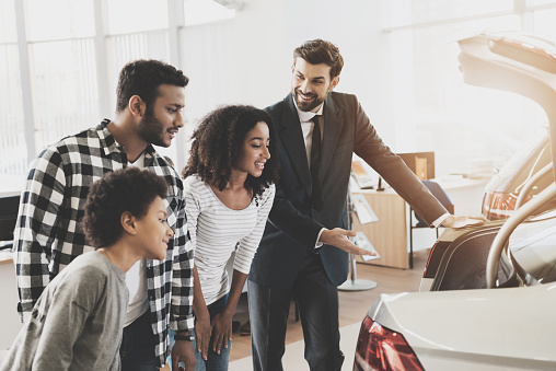 African American Family At Car Dealership. Salesman Is Showing Trunk Of New Car.