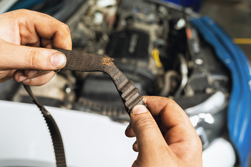 An Auto Mechanic Shows A Torn Timing Belt With Worn Teeth Against The Background Of An Open Car Hood Close Up
